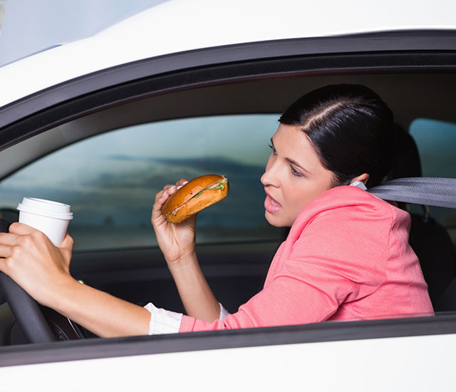 woman driving, eating her breakfast, holding a cup of coffee and talking on her cell phone